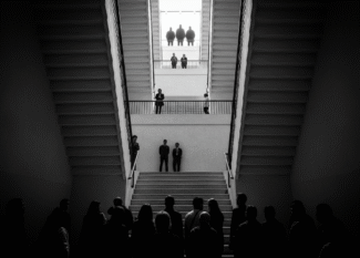 Foto em preto e branco de uma escadaria vista de baixo para cima, com silhuetas de pessoas em diferentes degraus (representa a hierarquia social)