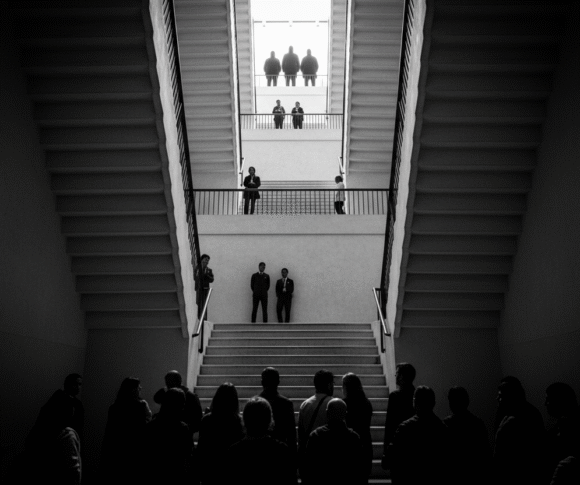 Foto em preto e branco de uma escadaria vista de baixo para cima, com silhuetas de pessoas em diferentes degraus (representa a hierarquia social)